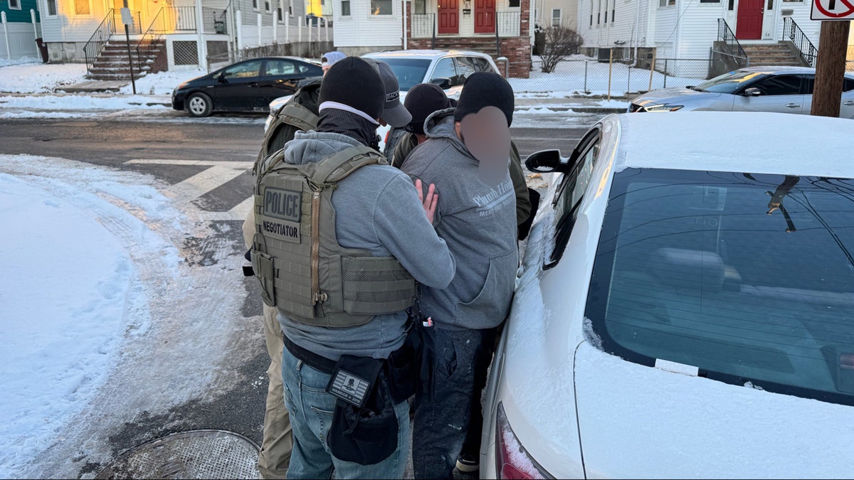 Three ICE officers stand around a man with a blurred out face as they put him in handcuffs.