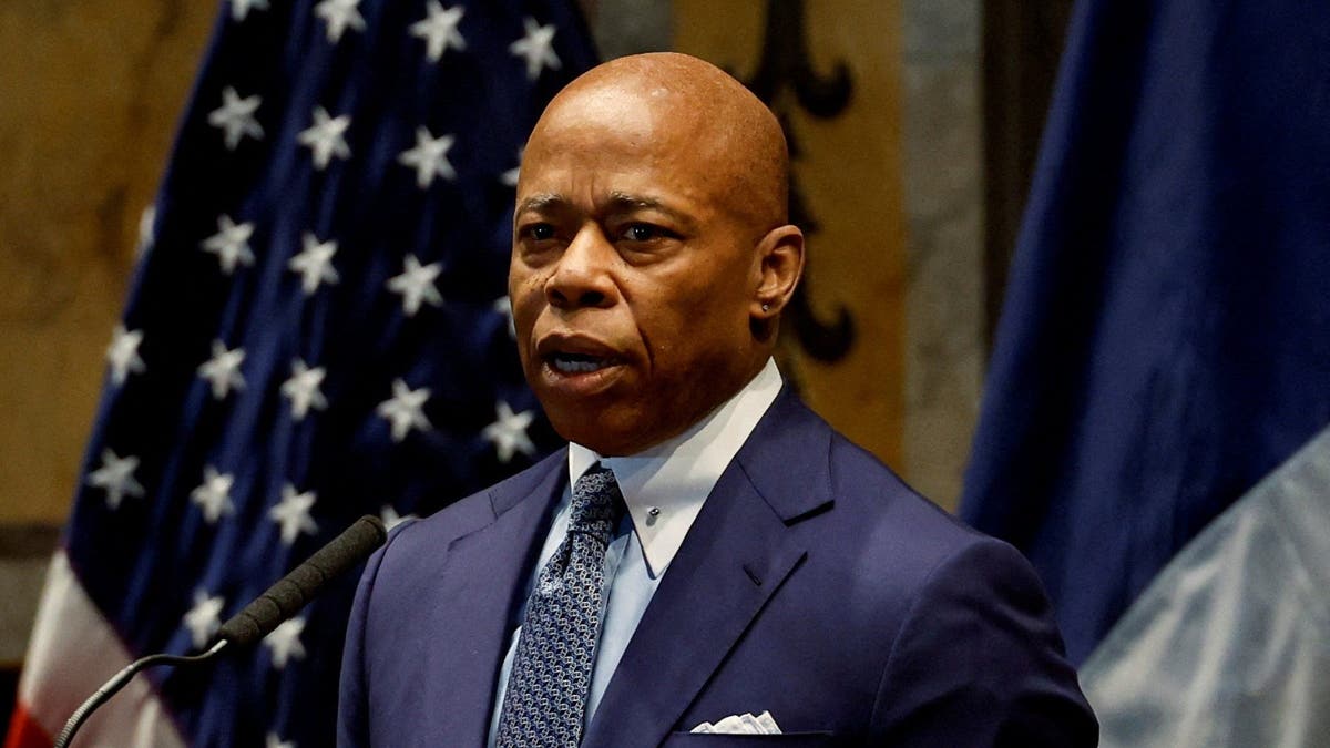 Eric Adams in a blue suit standing in front of a US flag while speaking