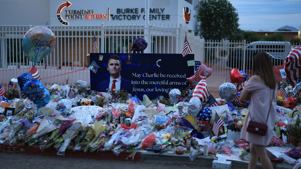 A woman in a pink dress approaches a makeshift memorial for Charlie Kirk outside a Turning Point USA office in Arizona at dusk