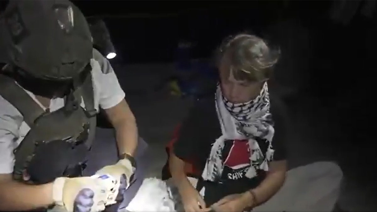 Greta Thunberg, wearing a black shirt and keffiyeh scarf, sits near an Israeli soldier in tactical gear.