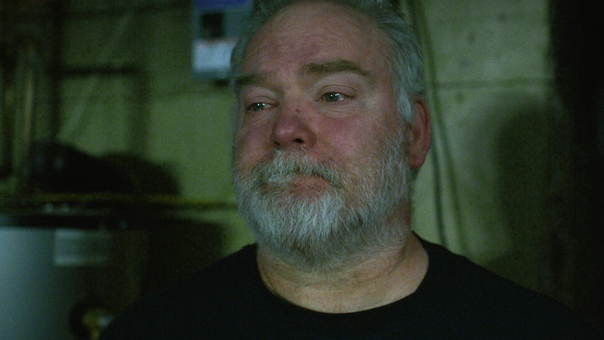 Mike Carroll stands in his dark basement with tears in his eyes and a sad expression.