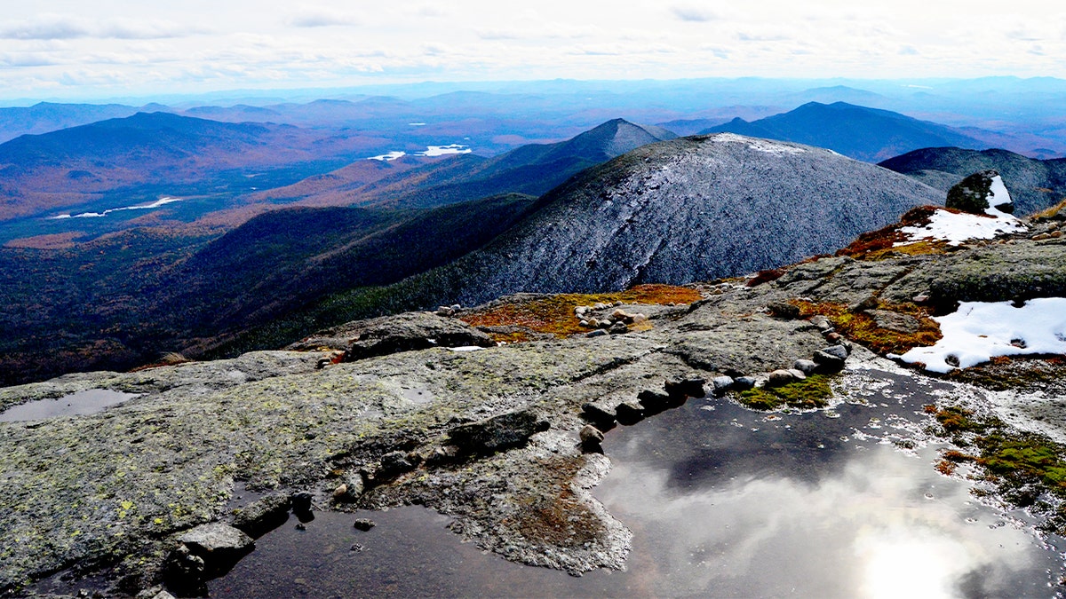 Expansive mountain panorama stretches in all directions from a rocky summit under a bright sky.