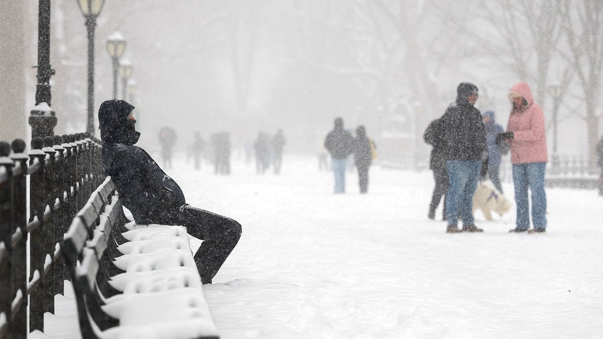 A bundled-up man rests on a park bench as snow falls steadily around him.