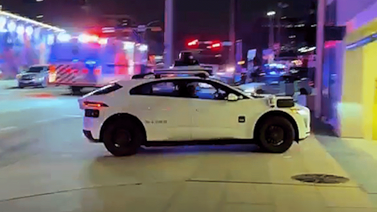 A Waymo autonomous vehicle drives through a brightly lit downtown Austin street at night near a parking garage entrance.