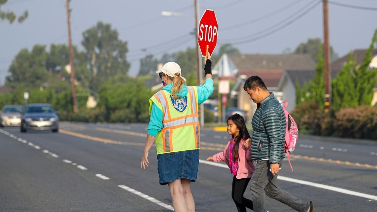 A crossing guard in a bright yellow vest helps a little girl dressed in pink and her father cross the road