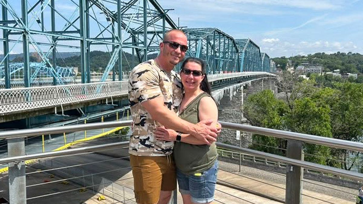 Justin Elrod and his wife, Lena Elrod, embracing while posing for a photo on an outdoor overlook with a steel truss bridge in the background.