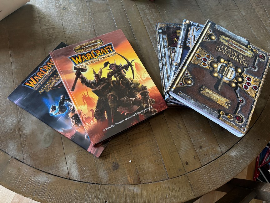 Two piles of books on a table: one for Warcraft's original D&D collaboration in the early 2000s, the other a stack of D&D 3.5 edition rulebooks that the Warcraft books were compatible with.