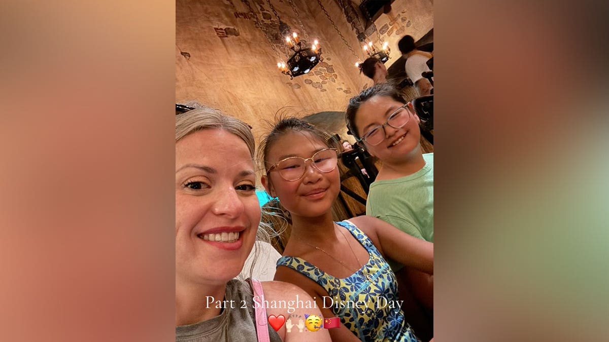 A woman takes a selfie with two young girls inside a dimly lit, stone-walled space with hanging chandeliers, with the overlaid text