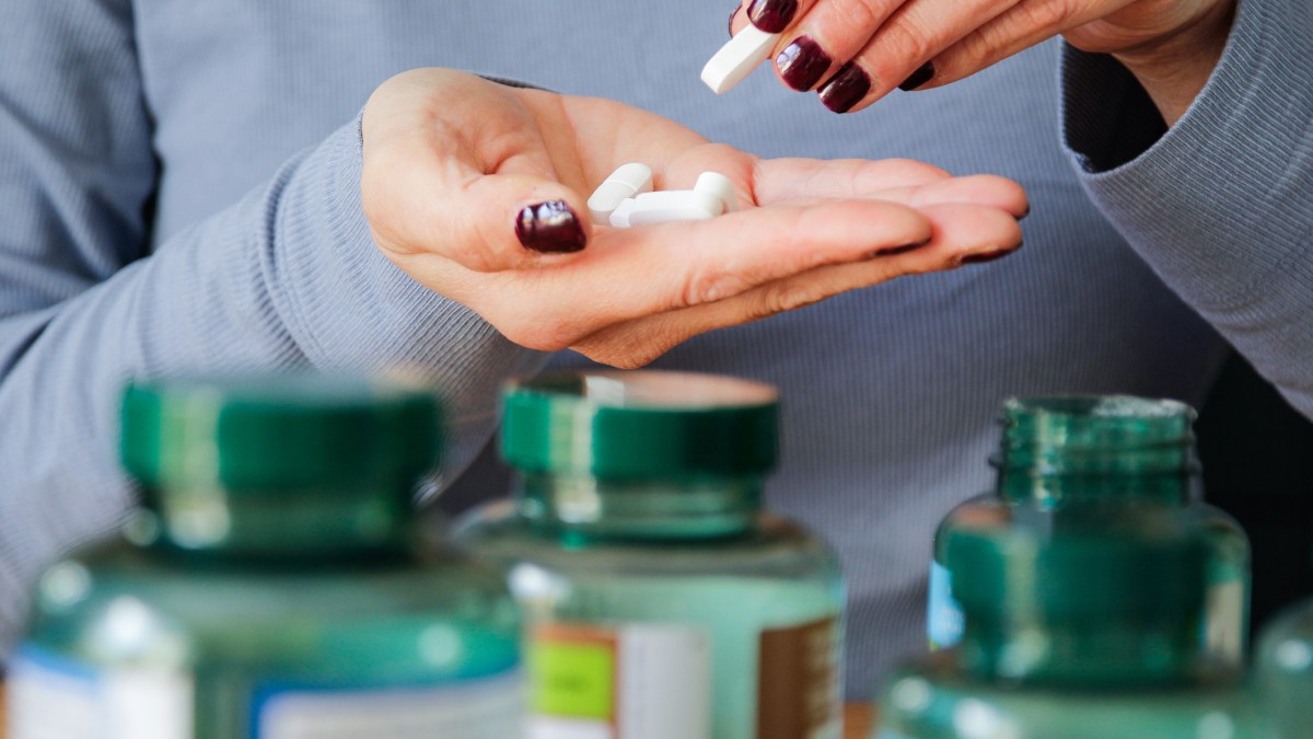 A person with dark red nails holding white pills in one hand with green vitamin bottles in the foreground.