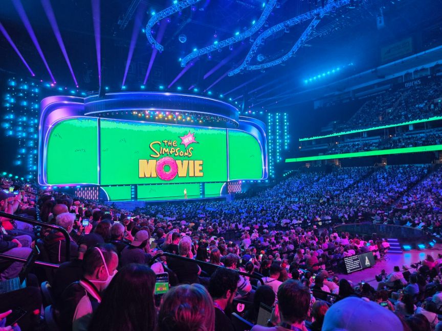 A large screen with the words "The Simpsons New Movie" in the animated series' iconic font in front of a large crowd of people.