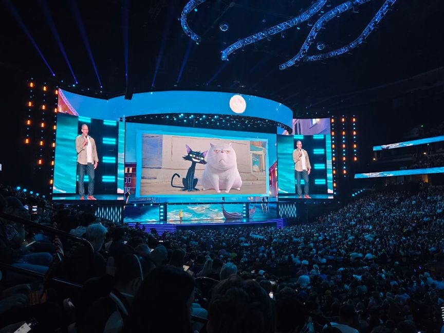 A man on stage talks in front of a screen with two animated cats on it in front of a crowd of people.