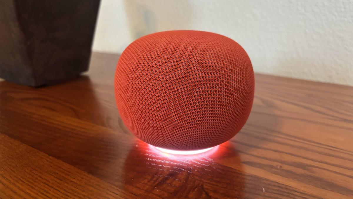 A red Google Home speaker on a wood desk next to a pot.