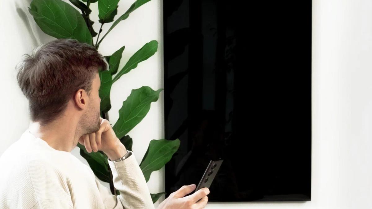 A man uses a remote on a black Kelvin heater on a white wall.