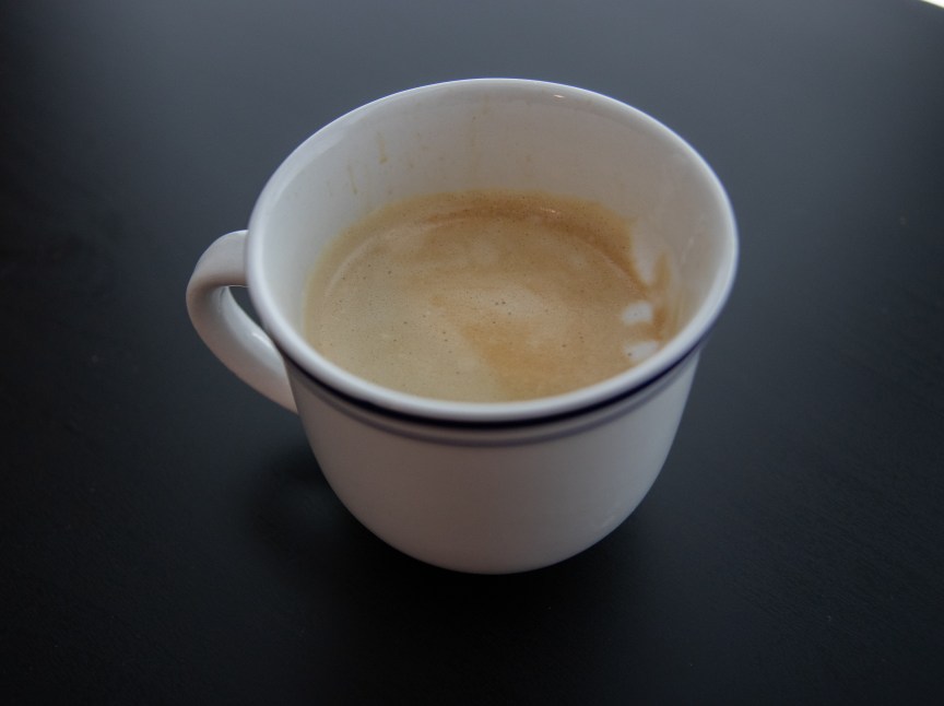 An espresso in a white ceramic cup, with a few drops of milk foam on top.