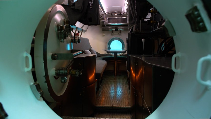 A round metal door opens into a living chamber featuring bunk beds, a meal table and a window looking out into a blue undersea world.