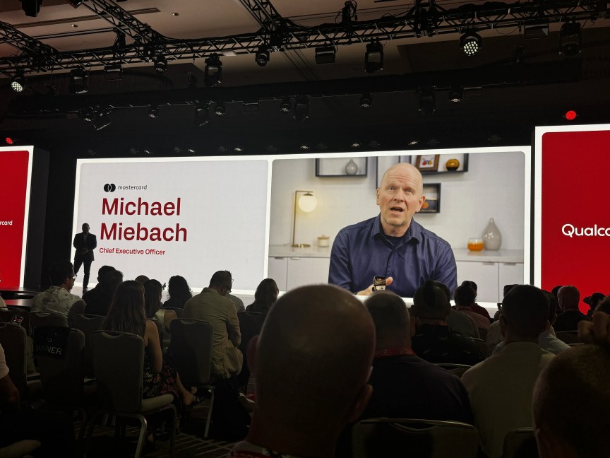 A man stands in shadow onstage while a prerecorded video plays behind him of an executive speaking.