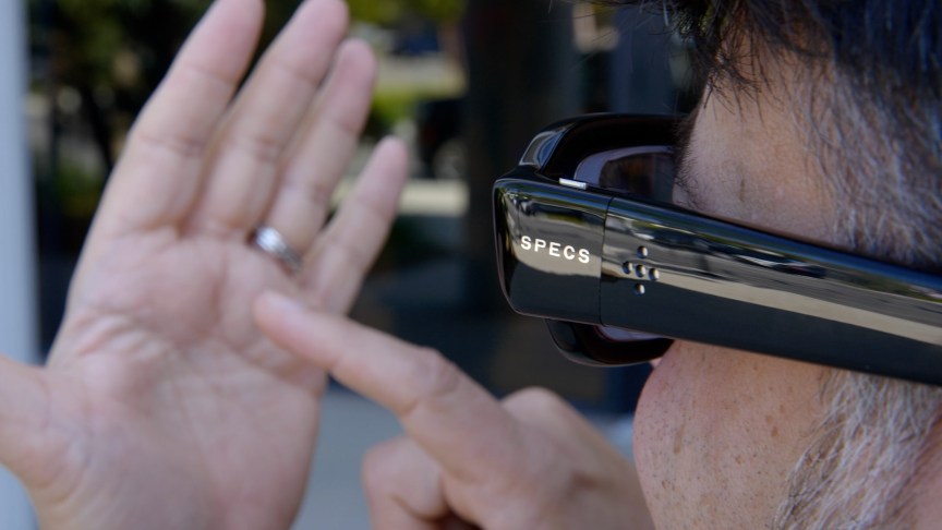 A hand raised up with a finger pointing at the open palm, while the person is looking through dark black AR glasses at it.